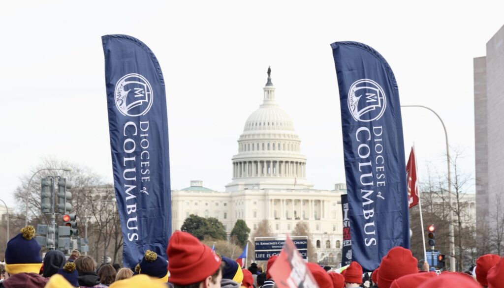 Crowd of people in red and yellow hats gathering along a street in front of the U.S. Capitol, with tall blue Diocese of Columbus banners visible on both sides.
