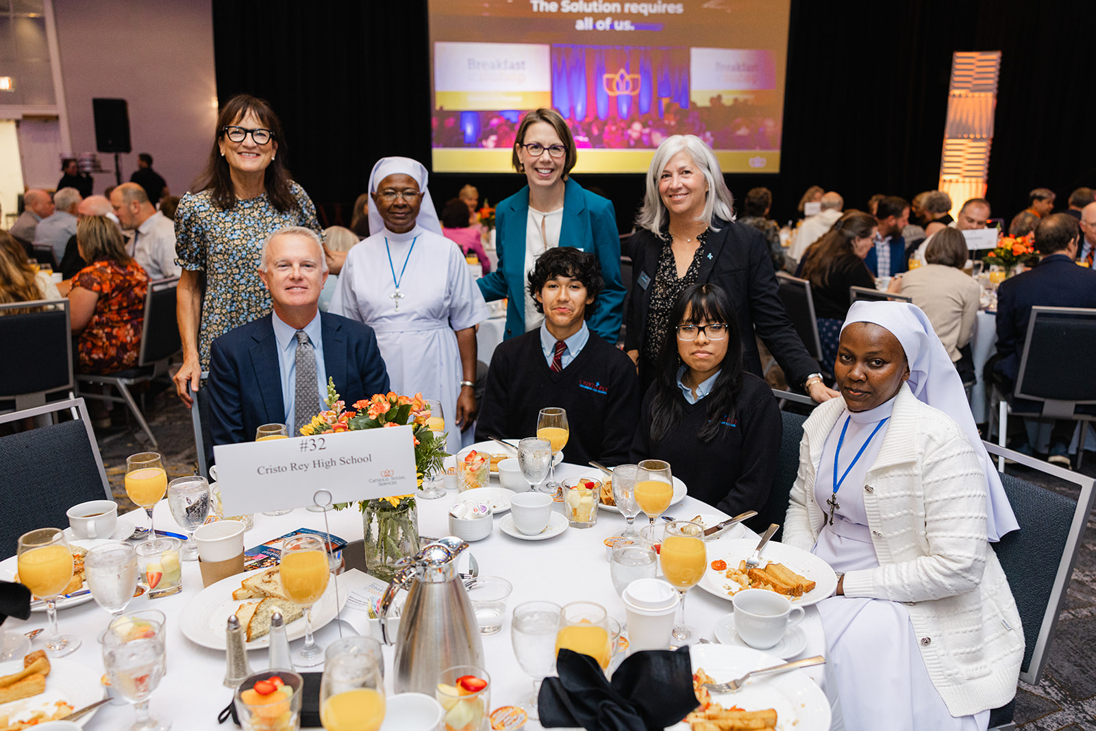 Group of eight people posing for a photo around a round banquet table at a formal event, with a nun and Cristo Rey High School sign on the table #32.