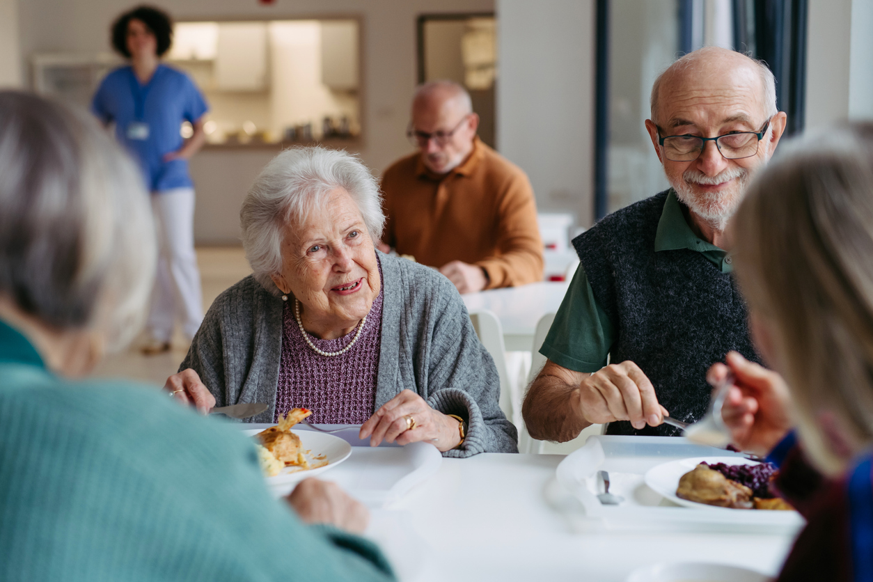 Group of seniors sharing a meal at a bright communal dining area, smiling and chatting at the table.