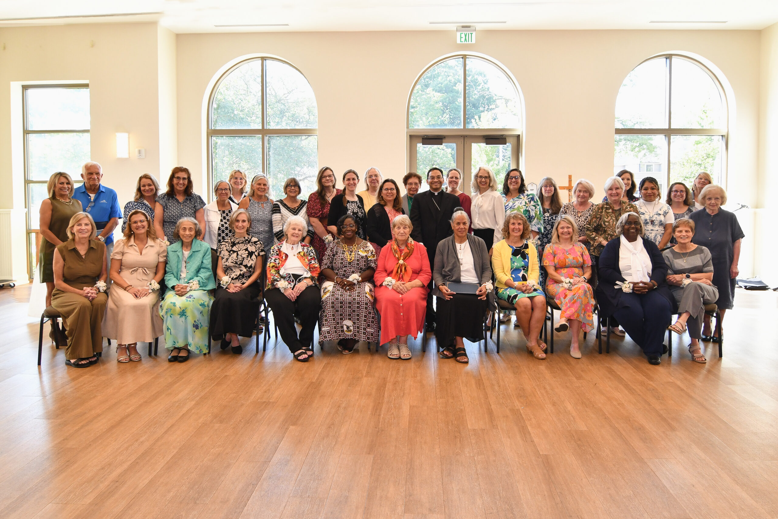 Group of people posing for a photo in a bright room with large arched windows, many seated in front and standing in back.