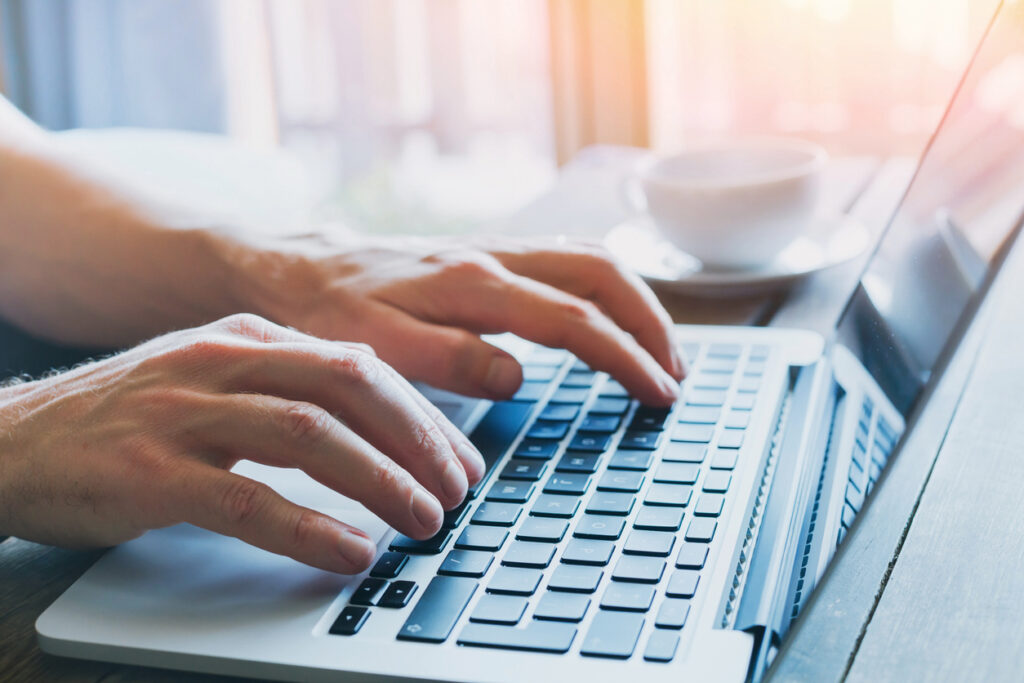 Close-up of hands typing on a laptop keyboard at a wooden desk with a cup of coffee in the softly lit background