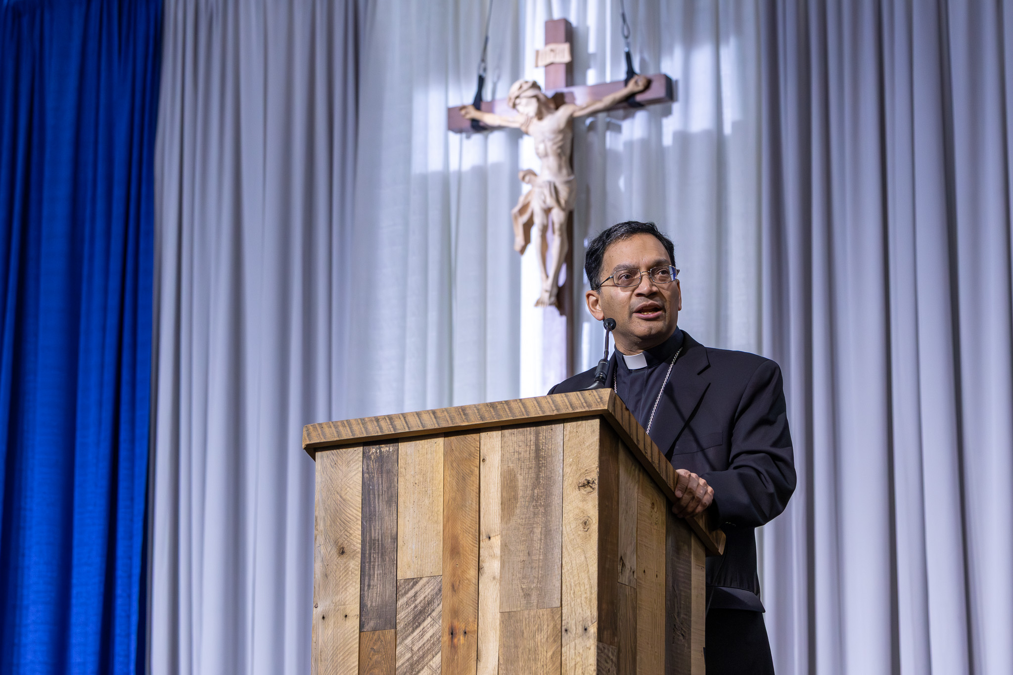 Clergyman in a black suit and clerical collar speaks at a wooden lectern with a crucifix behind him on a stage backdrop of blue curtains.